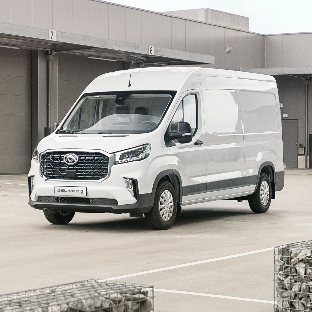 Weißer Lieferwagen DELIVER 9 auf einem Parkplatz, vor modernen Garagen im Hintergrund.