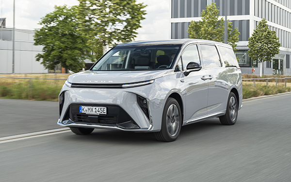 Ein silberner Elektro-Minivan fährt auf einer Straße, umgeben von Bäumen und modernen Gebäuden.